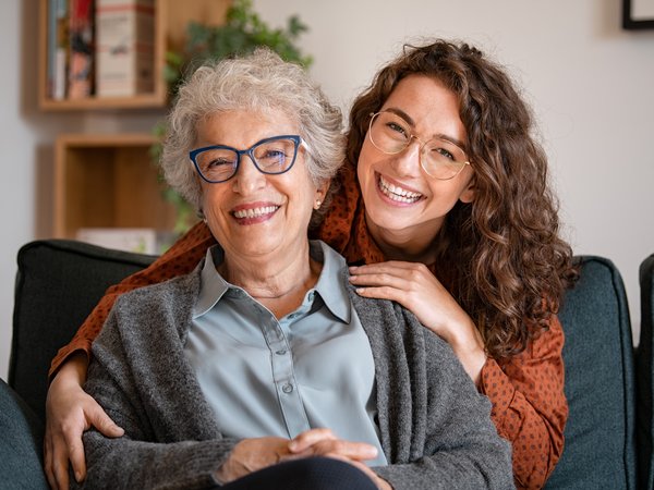 Glückliche Mutter mit lächelnder Tochter zu Hause. Lächelnde Mutter und Tochter sitzen gemeinsam auf dem Sofa in einem hellen Wohnzimmer und schauen glücklich in die Kamera.